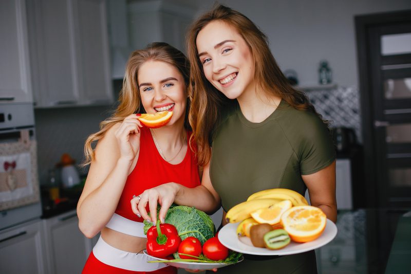dwie-sportsmenki-weganki-pozują-do-zdjęcia-w-kuchni-jedząc-zdrowe-warzywa